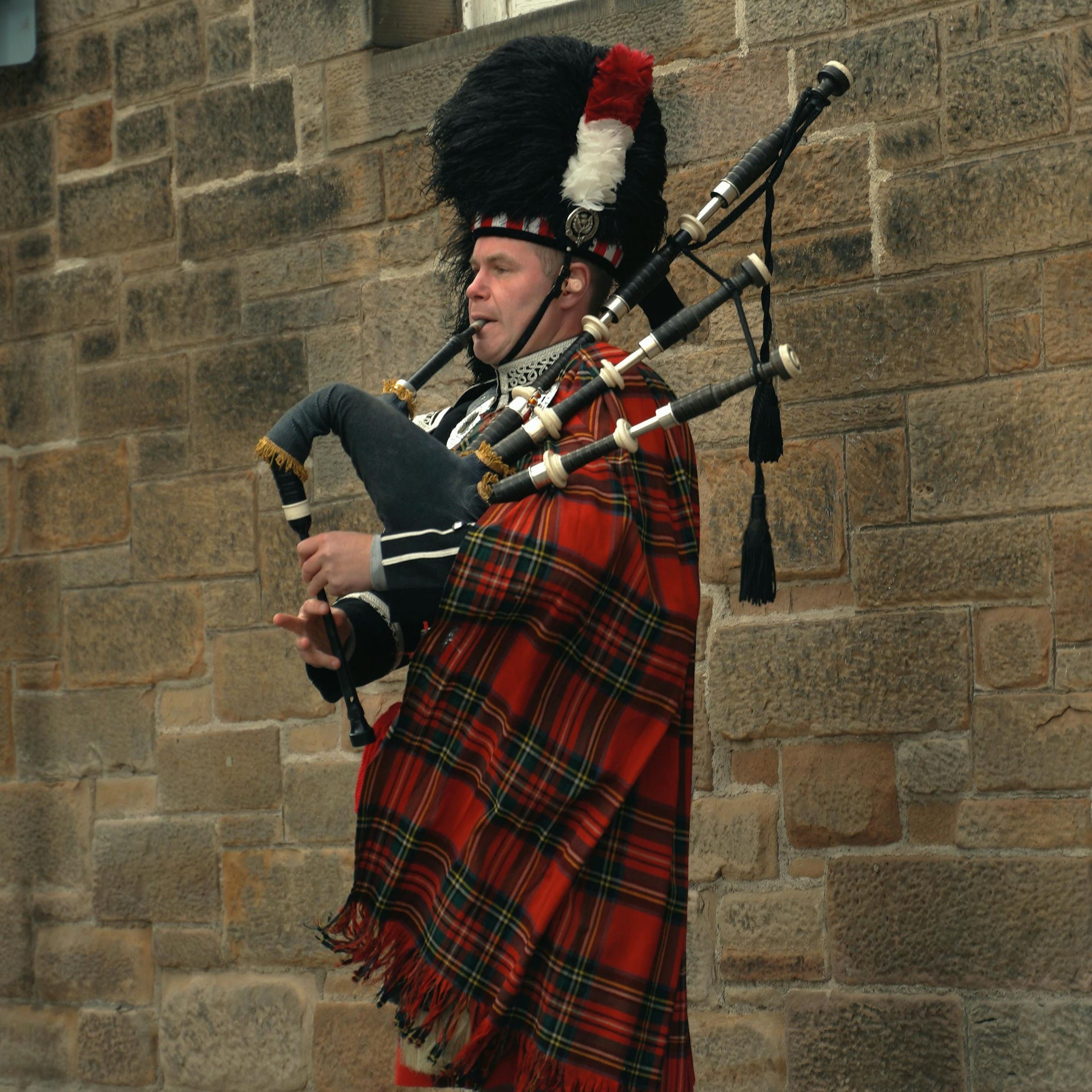 a person in traditional scottish attire plays the bagpipes. they wear a red tartan plaid and a black feathered hat, standing against a stone wall.