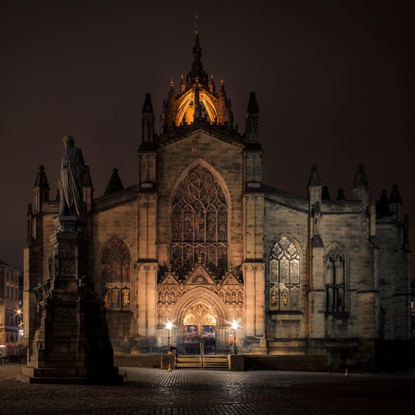 a grand, illuminated cathedral at night, with intricate gothic architecture and glowing stained glass windows. a statue stands on a tall pedestal in front of the church, casting a shadow on the cobblestone plaza.