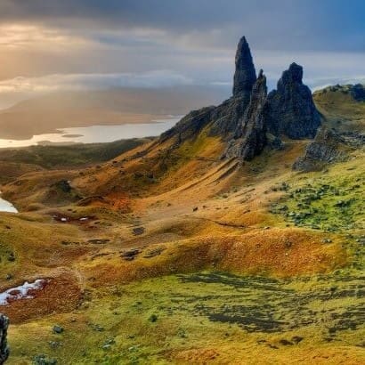 a dramatic landscape featuring the old man of storr rock formation on the isle of skye, scotland. the scene shows rugged, green hills with patches of orange and brown under a cloudy sky, with sunlight breaking through in the distance.