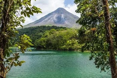 a distant volcanic mountain peak is framed by lush green trees and foliage. in the foreground, a serene body of water reflects the vibrant greenery. the sky is partly cloudy, adding depth to the landscape.