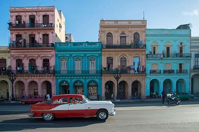 a red and white classic car cruises by colorful, historic buildings in vibrant shades of pink, blue, and turquoise under the cuban sun. a few people and motorcycles add to the lively scene in this dream destination.