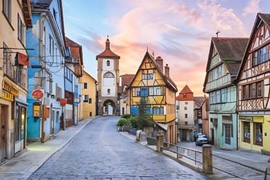 a picturesque street in a quaint european town, possibly a hidden gem of germany, features colorful, half-timbered houses and a cobblestone path leading to an arched tower. this idyllic destination is bathed in vibrant pink and orange hues at sunset, perfect for any travel enthusiast.