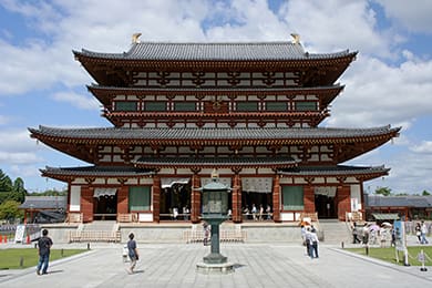 under a partly cloudy sky, travelers explore a large, traditional japanese temple. the intricate wooden architecture and multiple tiers captivate tourists as they wander through the stone courtyard in front of this iconic gem in japan.