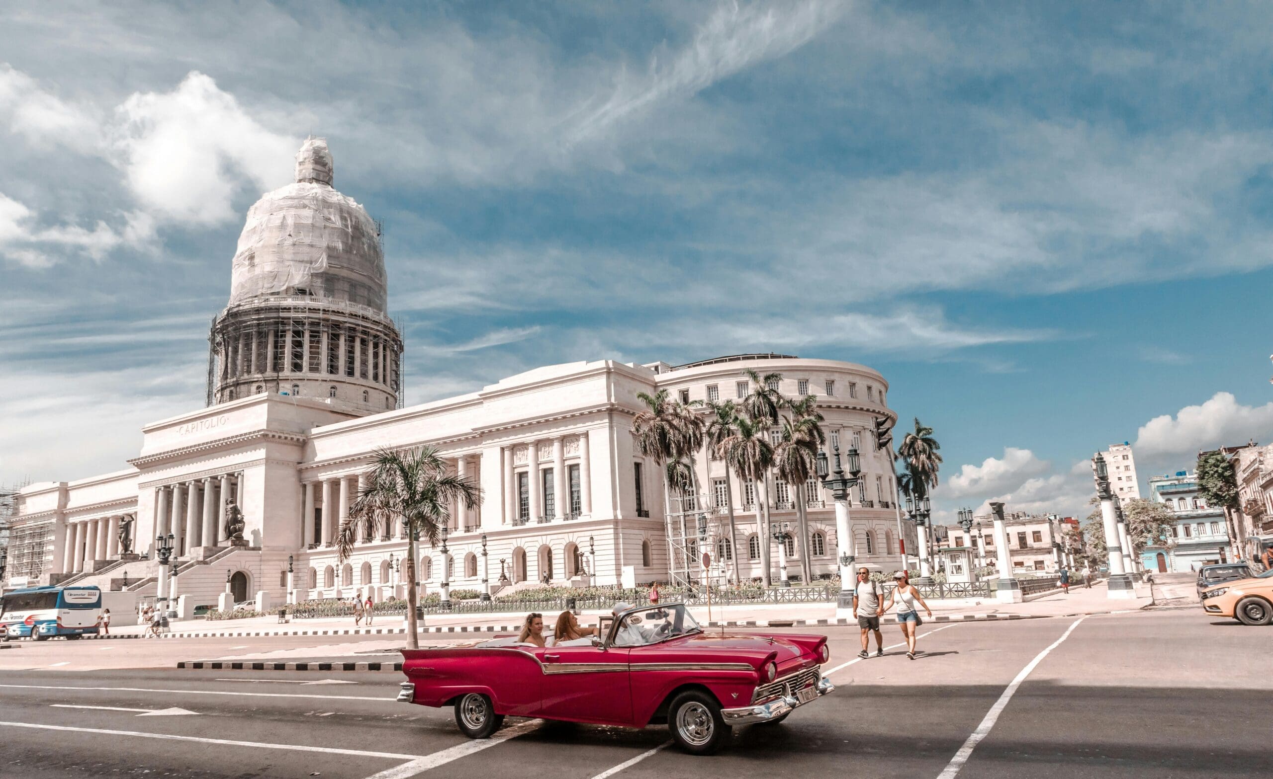 a vintage red convertible with people inside drives past a large, ornate building resembling a capitol. the sky is blue with scattered clouds, and palm trees line the street, creating a picturesque, sunny scene.