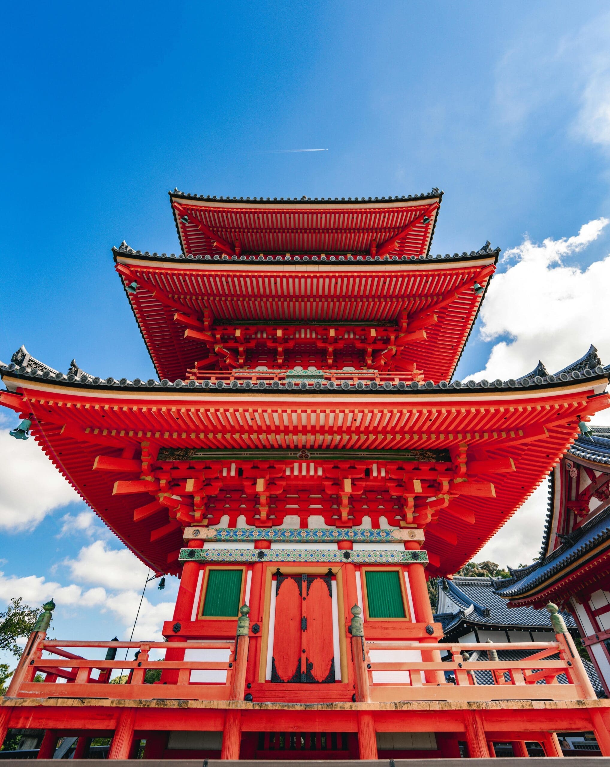 a vibrant red, multi-tiered pagoda with ornate wooden details stands against a bright blue sky. wispy clouds and a contrail are visible in the background, enhancing the structure's majestic appearance.